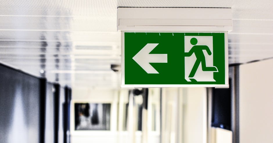 A green emergency exit sign with a white running figure and an arrow pointing left, mounted on a white ceiling in a corridor. The corridor features white panels and appears to be inside a building, potentially in a residential or commercial property. The sign indicates the direction for evacuation, which is relevant in emergency situations, and is positioned above a passageway or doorway. The area is well-lit with natural or artificial lighting, supporting safety measures during home relocations or last-minute moves. Man and a Van Paddington may utilize such signage during the process of house removals, ensuring safe and efficient navigation for movers and clients in the W2, Paddington area, especially in urgent or emergency evacuation scenarios involving furniture transport and packing logistics.