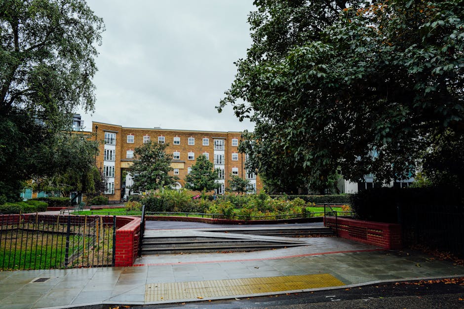The image shows a residential street scene with an entrance to a garden area, enclosed by a black metal fence with red brick columns on either side. The entrance has a set of steps leading up to a paved pathway, which is partially covered with a black protective blanket or moving pad, indicating a furniture transport or home relocation process. Surrounding the garden are well-maintained flower beds and greenery, with several mature trees providing partial shade. In the background, there is a multi-storey brick apartment building with white-framed windows and balconies, typical of London’s housing architecture. The sky overhead is cloudy and gray, suggesting overcast weather. This scene reflects a typical setting for a house removal service, such as those provided by Man and a Van Paddington, involving the careful handling and transport of furniture and belongings during a move into or out of the residential building, with focus on the logistics of loading or unloading at the property’s entrance.