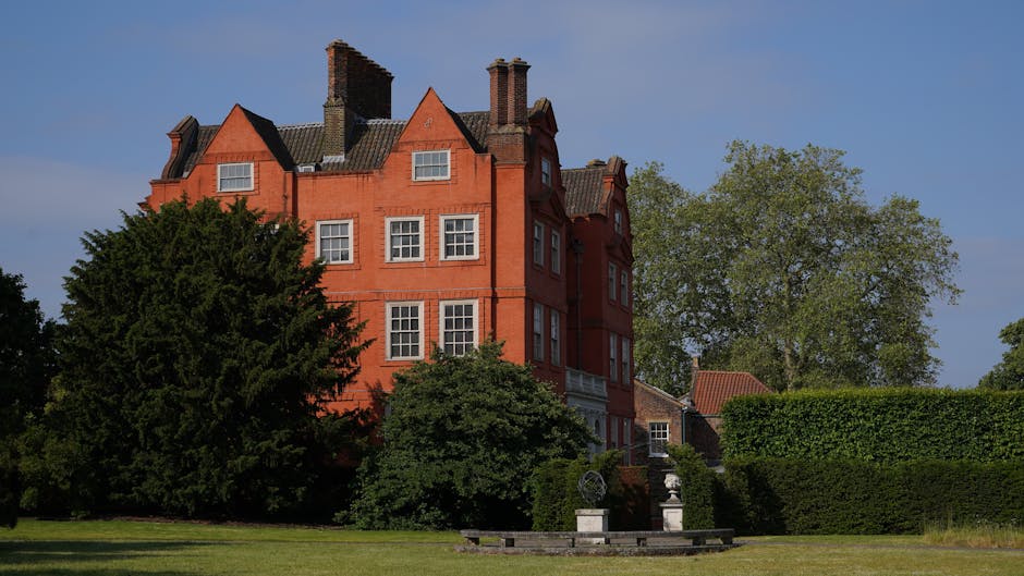 A large red-brick residential building with multiple carved stone chimneys and dormer windows, situated behind a landscaped garden area with lush green trees, including a tall conifer and a deciduous tree. In the foreground, there is an open lawn with a stone pathway and a decorative stone fountain. A man from Man and a Van Paddington is engaged in a home relocation process outside the property, managing cardboard boxes and wrapped furniture items near the building's entrance, with visible packing materials such as plastic wrap and blankets. A moving van is parked nearby, indicating the loading and furniture transport process involved in house removals and packing for the move to Norfolk Square Gardens Residences, Paddington.