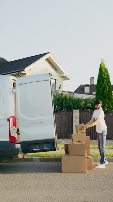 A person standing on a paved street outside a residential property, engaged in the home relocation process, with the rear door of a white moving van open. The individual is wearing casual clothing and