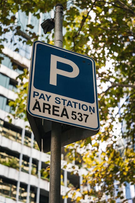 A blue and white parking sign attached to a metal pole, indicating a pay station area with the number 537, located outdoors on a street with surrounding trees and modern office buildings in the backgr