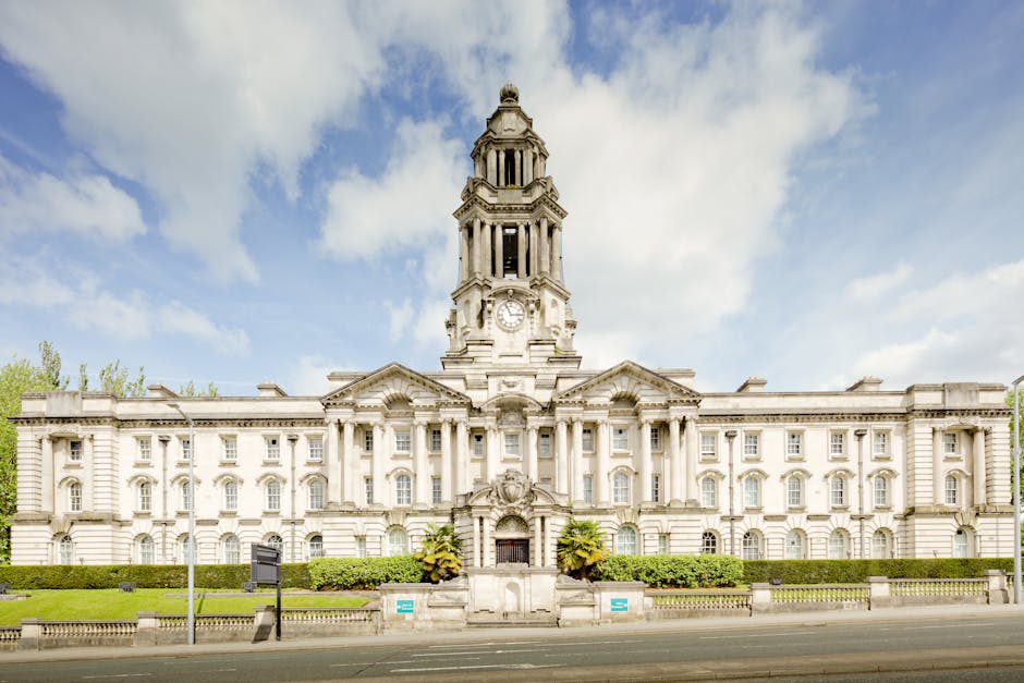 A large historic building with classical architectural features, including a prominent central clock tower topped with a decorative spire. The facade is constructed from light-colored stone with evenl